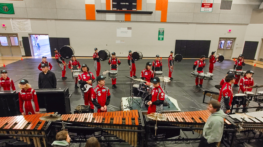 Indoor percussion ensemble performing with marimbas and marching drums in a gymnasium