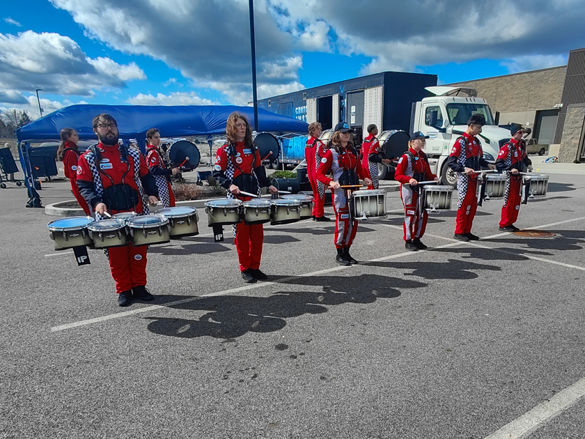 Marching band drumline in red uniforms rehearsing outdoors with snare and tenor drums