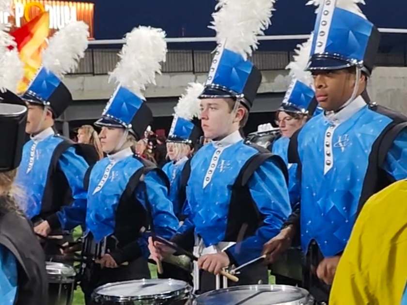 Marching snare line in blue geometric uniforms with white plumes performing at a night stadium show