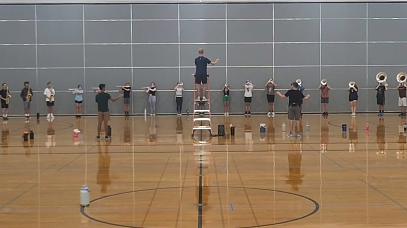 Marching band winds rehearsing indoors while a director conducts from a ladder